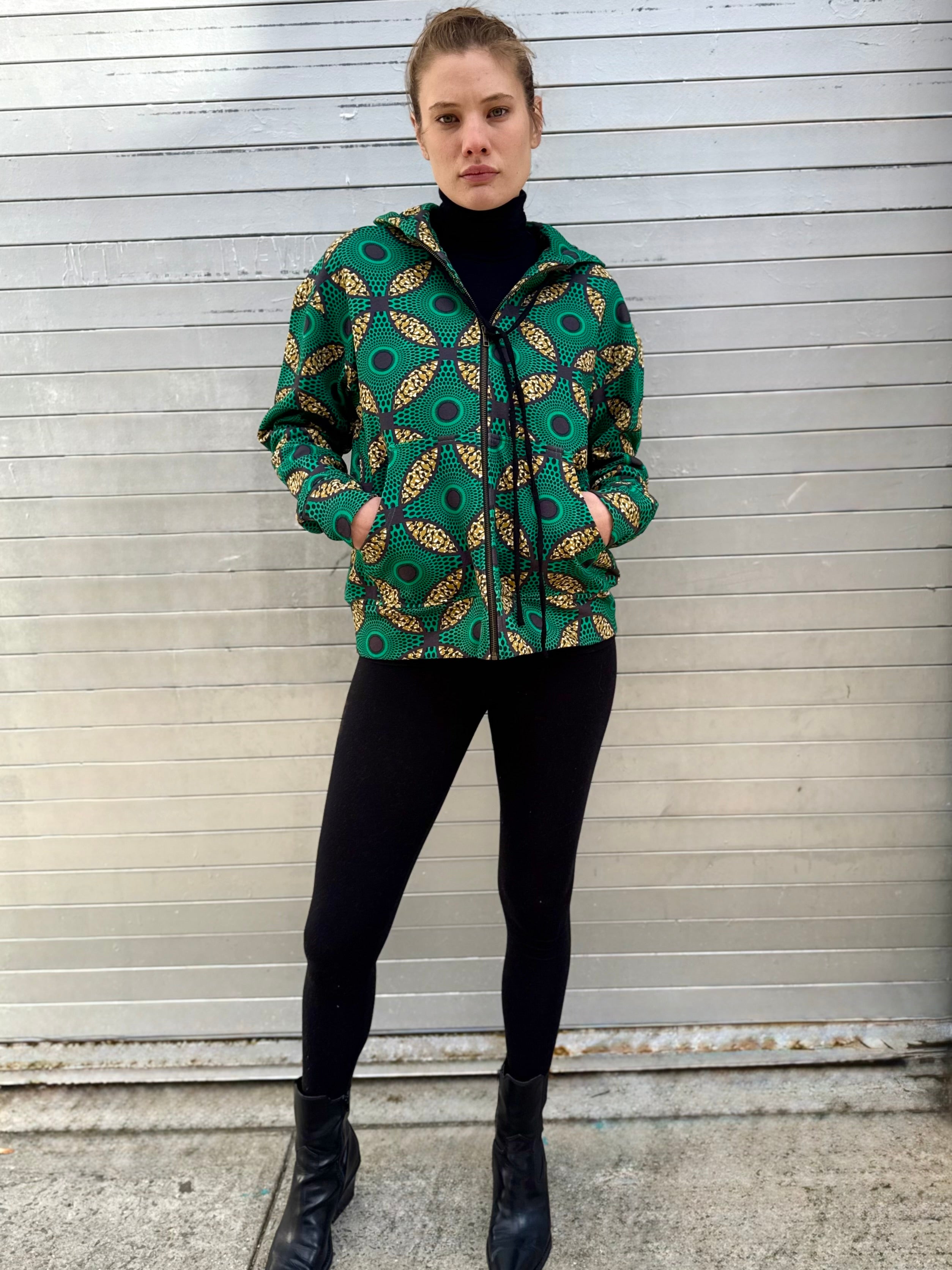 Full-body view of model wearing the Ìmọ́lẹ̀ Green Hoodie with black pants and boots. Crafted from organic cotton with eco-printed Yoruba patterns, this sustainable zip-up represents the fusion of bold design, cultural storytelling, and everyday comfort.