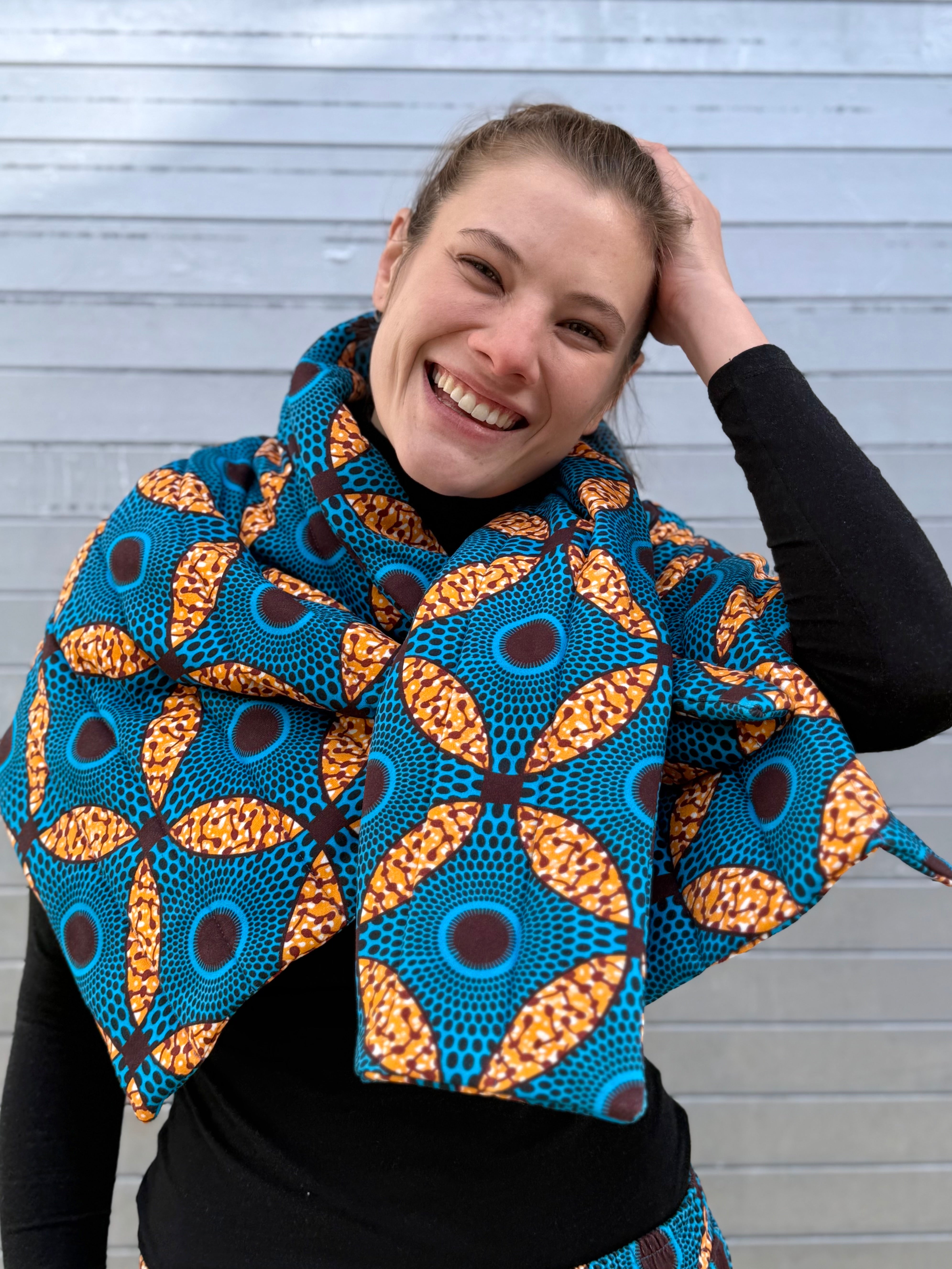 Model smiling while wearing the Ìmọ́lẹ̀ Padded Sweat Scarf in blue and orange African print by Lola Faturoti Loves. A joyful expression of color and culture: the Ìmọ́lẹ̀ Scarf celebrates empowerment through sustainable design.
